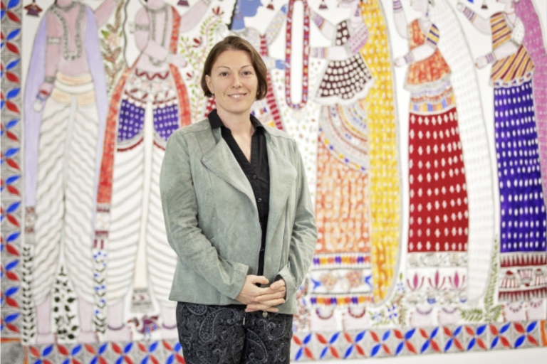 A woman stands indoors in front of a colorful, patterned mural featuring stylized figures in traditional attire.