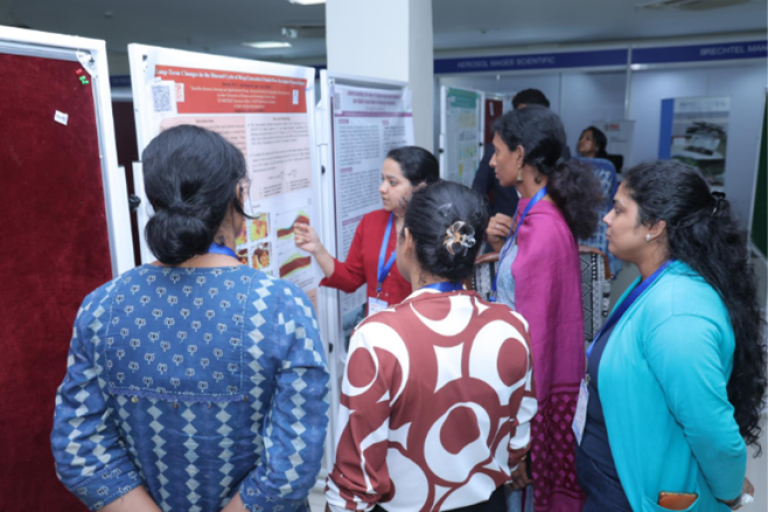 A group of women stand around a presenter explaining information displayed on a scientific poster at an indoor event or conference.