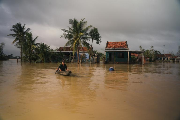 A person paddles a small boat through floodwaters in front of partially submerged houses and palm trees under a cloudy sky.