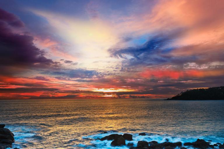 Colorful sunset over the ocean with waves crashing against rocks in the foreground and partly cloudy sky filled with vibrant hues of orange, pink, blue, and purple.