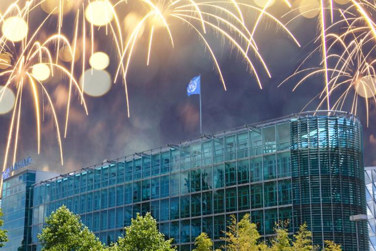 A modern glass office building with a blue flag on the roof, surrounded by trees, with fireworks lighting up the night sky above.
