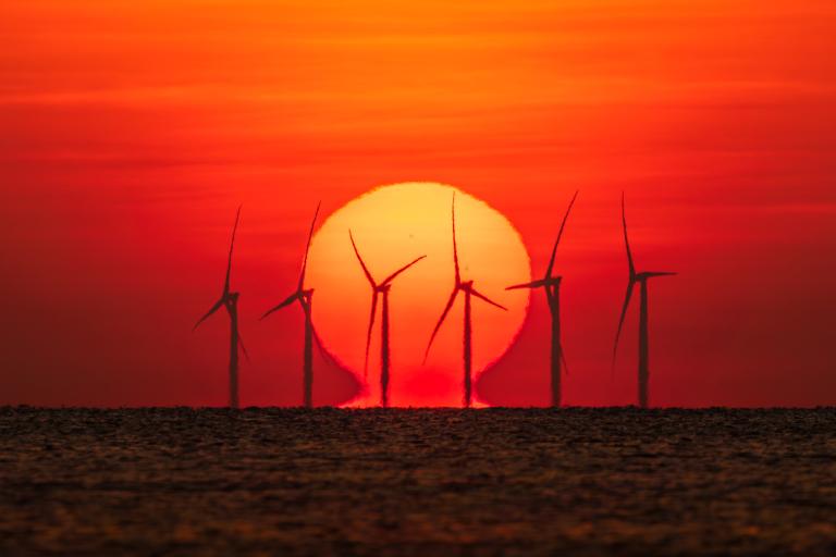 Wind turbines are silhouetted against a large setting sun over the ocean, with the sky glowing in shades of orange and red.