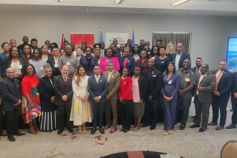 A large group of professionally dressed people pose for a group photo in a conference room, with banners in the background.