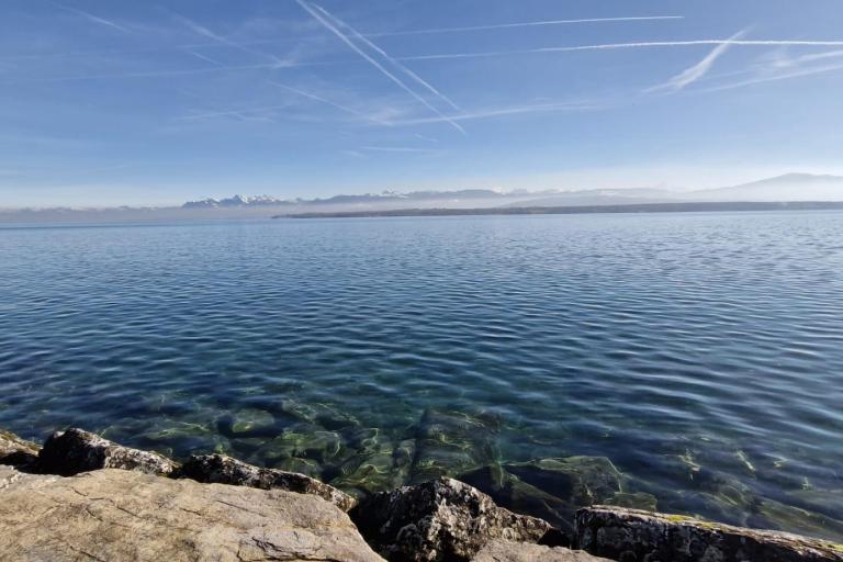 Clear blue lake with visible rocks beneath the water in the foreground, distant mountains on the horizon, and several contrails in the sky.