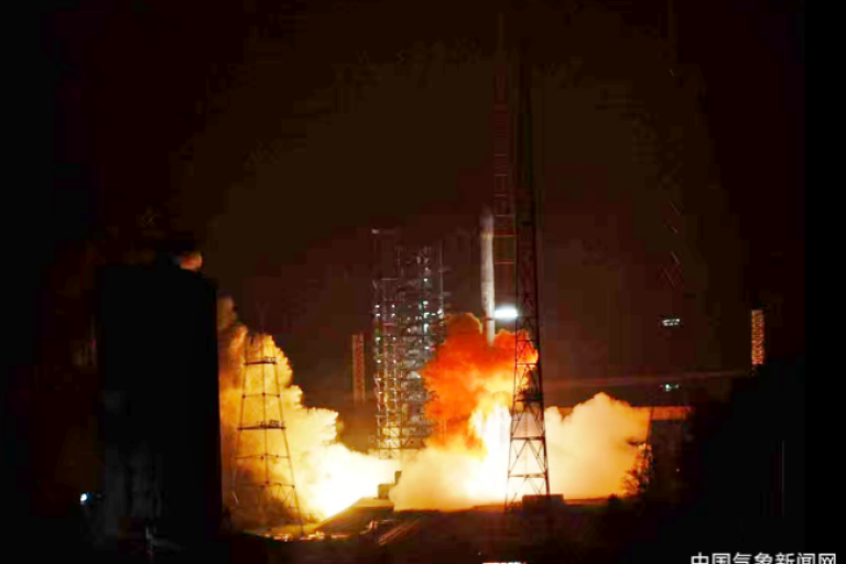 A rocket launches at night, emitting bright flames and smoke, with metal structures and a lit-up tower visible in the background. Chinese text is present in the bottom right corner.