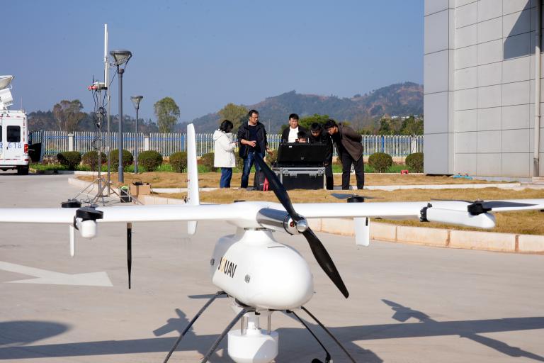 Four people stand by a black equipment case outdoors while a large white drone marked “XAMY” rests on the pavement in the foreground.