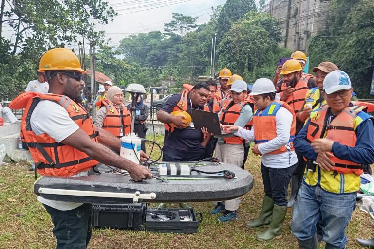 A group of people wearing safety helmets and life vests gather outdoors around equipment, with one person using a laptop and another handling a survey instrument.
