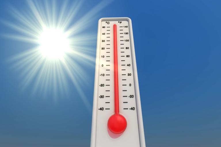 A thermometer showing a high temperature stands against a clear blue sky with the sun shining brightly in the background.
