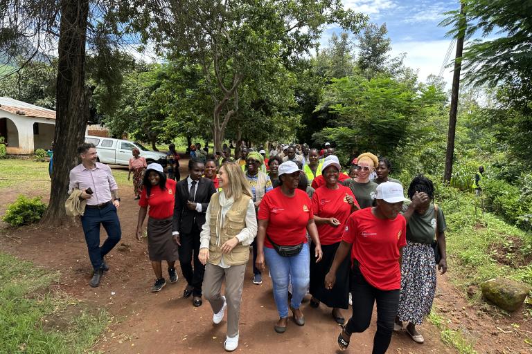 A group of people, including women in red shirts and white caps, walk together outside on a tree-lined path, with others and a white van visible in the background.