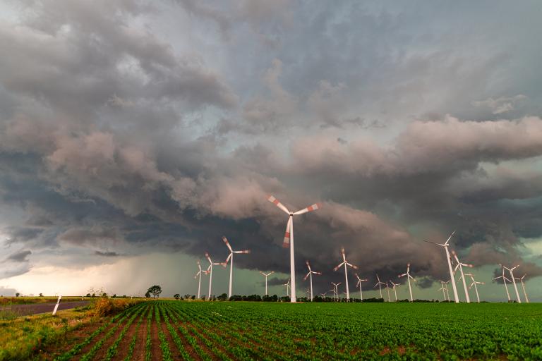 Wind turbines in a field with storm clouds overhead, green crops in the foreground, and dramatic sky in the background.