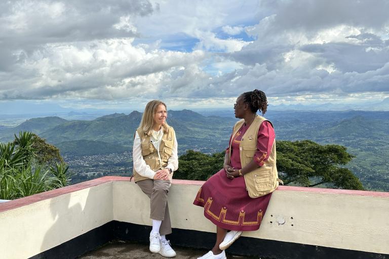 Two women sit on a low wall overlooking a scenic mountain landscape under a cloudy sky. Both wear beige vests; one is in a dress, the other in pants and a jacket.