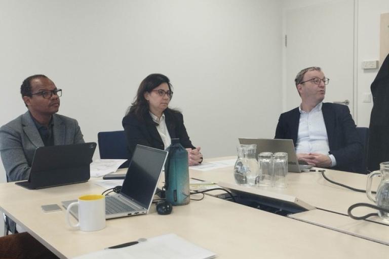 Three people sit at a conference table with laptops, notebooks, a water bottle, and glasses, appearing to attend a meeting in a modern office setting.
