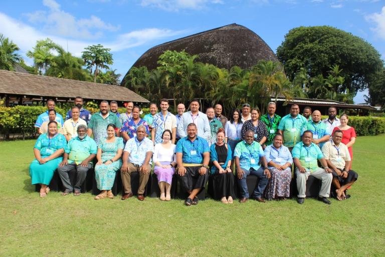 A large group of adults poses for a photo on grass in front of a tropical building with thatched roof and palm trees, under a partly cloudy sky.