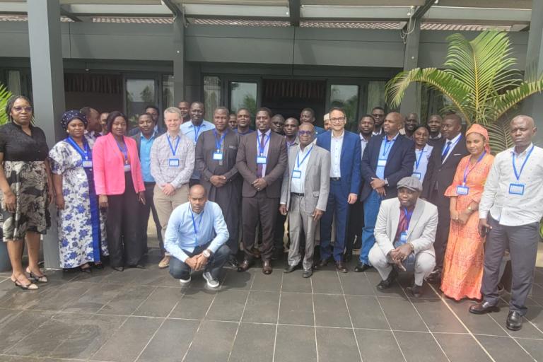 A group of people wearing conference badges pose for a group photo outdoors on a tiled surface, with a building and a potted plant in the background.