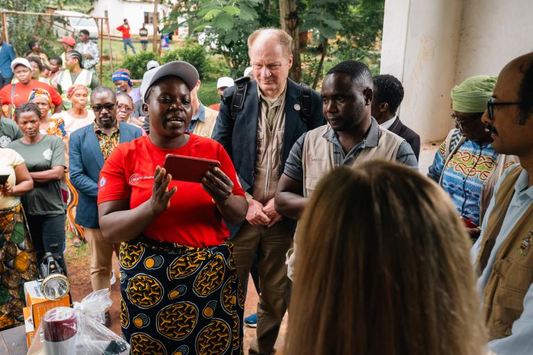 A woman in a red shirt speaks to a group of people gathered outdoors, some wearing badges and vests, while others observe in the background.