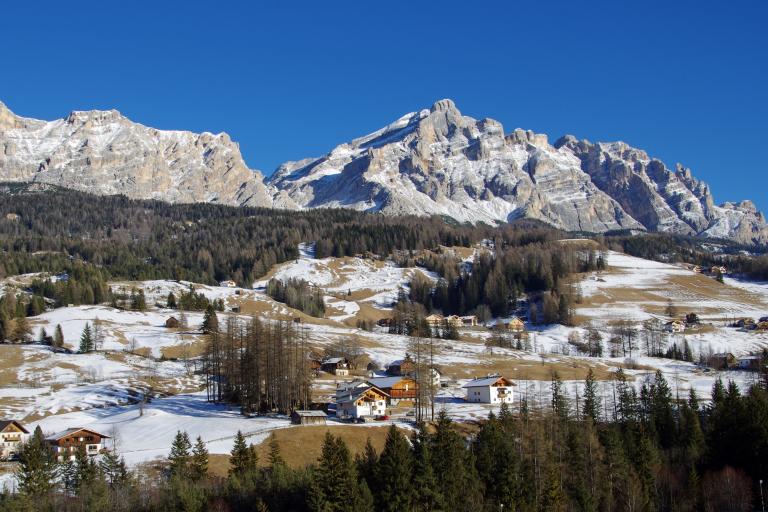 Snow-dusted mountains and scattered houses on a hillside under a clear blue sky, with patches of trees and grass visible.
