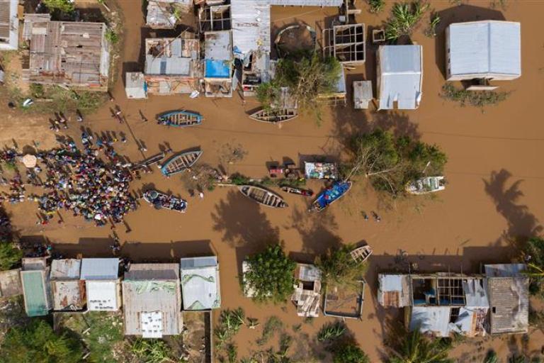 Aerial view of a flooded village with submerged houses, boats, and a large group of people gathered on a dry patch of ground.