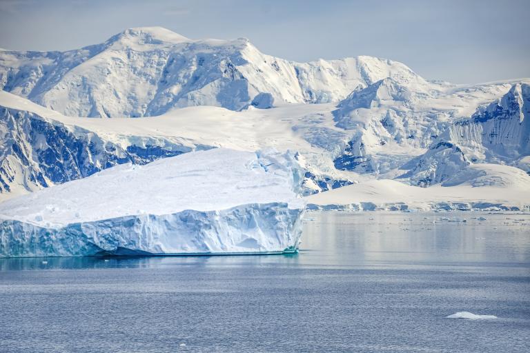 Large iceberg floating in calm water with snow-covered mountains in the background under a cloudy sky.