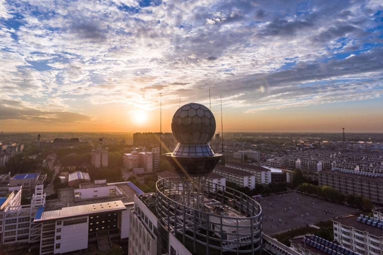 A cityscape at sunset featuring a spherical structure atop a rooftop, with buildings and a cloudy sky in the background.