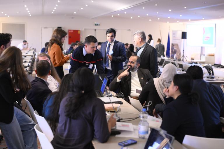 A group of people in business attire are gathered around tables with laptops, engaged in discussion at a conference or workshop in a well-lit room.