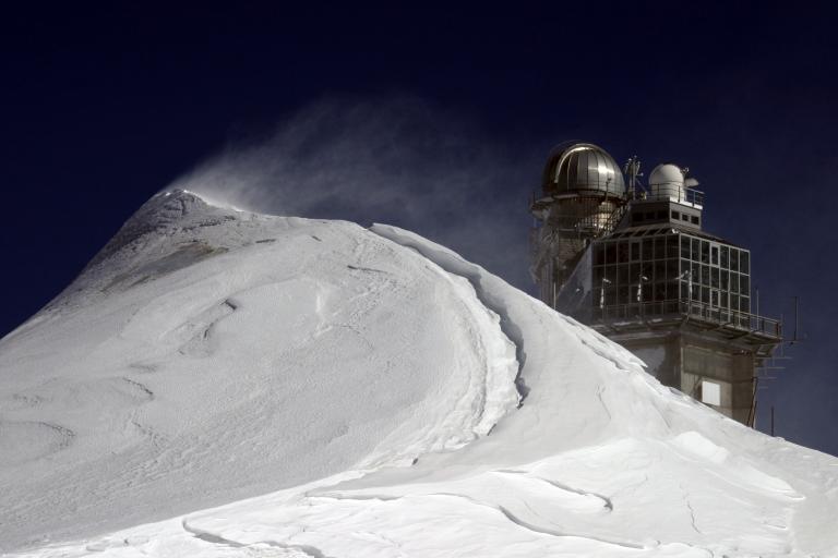 Snow-covered peak with wind-blown snow in the foreground and a building with a dome observatory structure in the background against a dark sky.