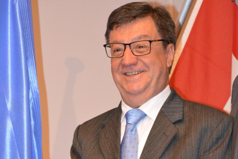 A man in a suit and glasses smiles while standing in front of blue and red flags.