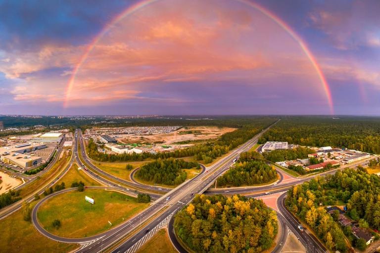 Aerial view of a highway interchange surrounded by greenery with a vivid rainbow arching across a partly cloudy sky at sunset.