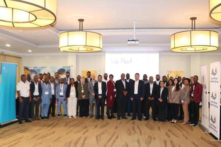 A large group of people pose for a photo in a conference room with banners and presentation screens in the background.
