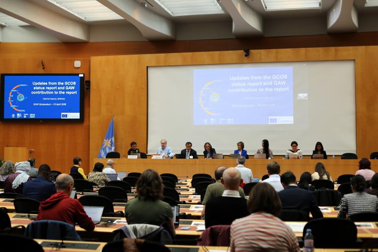 A panel of eight people sits at a conference table in front of an audience, with presentation slides displayed on large screens behind them in a modern meeting room.
