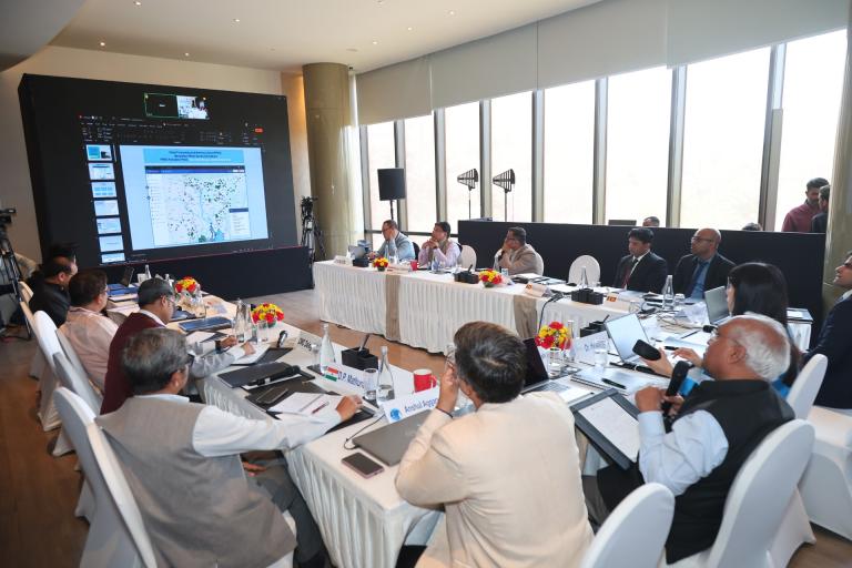 A group of people in formal attire sit around a U-shaped table with laptops and documents, viewing a large screen displaying a map and data in a modern conference room.