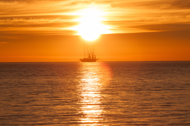 A tall ship sails on calm water at sunset, silhouetted against the bright orange sky with the sun low on the horizon and its reflection on the sea.