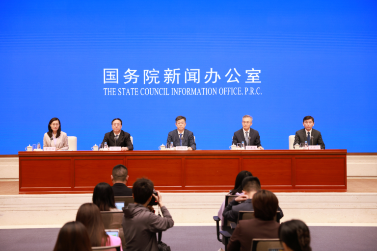 Five officials sit at a long table during a press conference, with "The State Council Information Office, P.R.C." displayed in English and Chinese on a blue screen behind them.