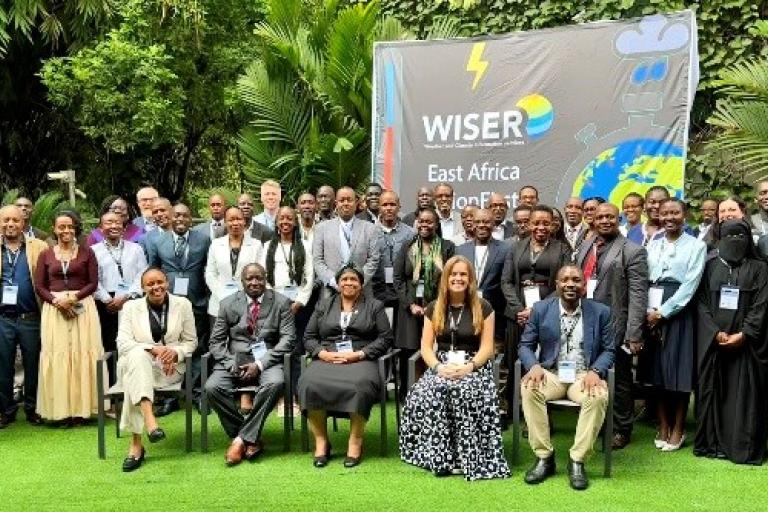 A large group of people pose for a photo outdoors in front of a sign that reads "WISER East Africa," surrounded by greenery.