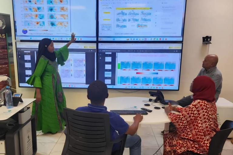 Four people in an office look at large screens displaying maps and data charts; one woman stands and points to the screen while others sit and observe.