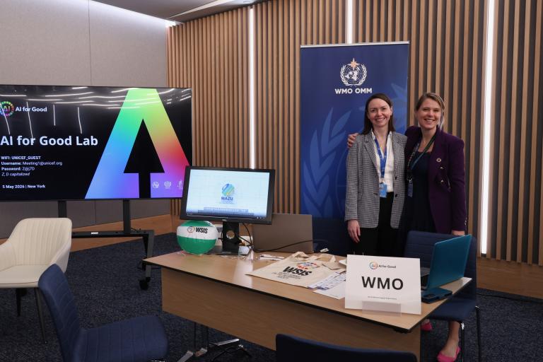 Two women stand smiling behind a desk with a "WMO" sign, in an office with a large screen displaying "AI for Good Lab" and WMO logo on the wall.