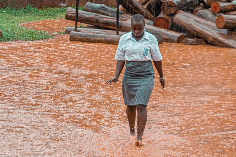 A student in uniform walks barefoot through a flooded, muddy area during heavy rain, with stacked logs in the background.