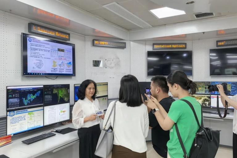 A woman stands in front of several monitors displaying weather data as she is interviewed and photographed by a group of people in a control room.