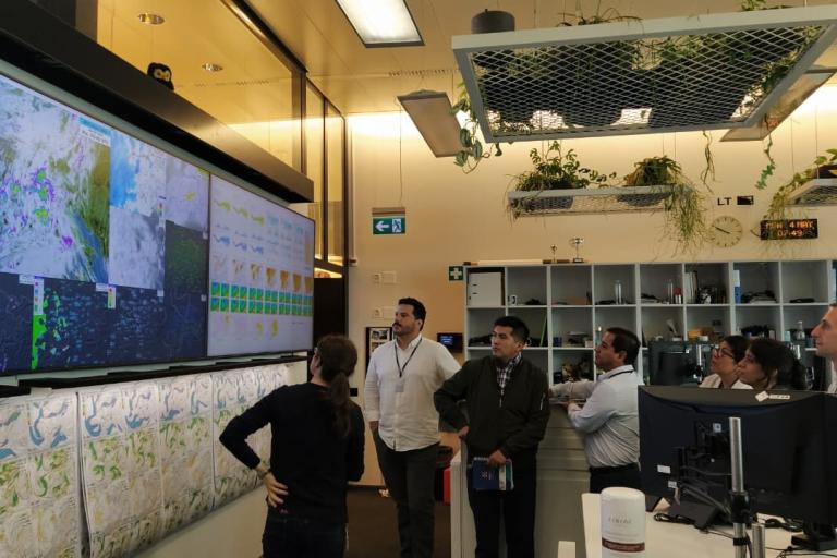 A group of people stand in front of large screens displaying weather maps and data in a modern office with plants and shelves in the background.