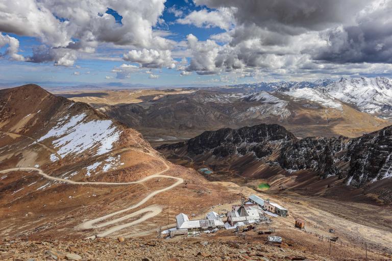 Mountain landscape with a winding road leading to buildings at the base, surrounded by rocky hills, snow patches, and distant snow-capped peaks under a cloudy sky.