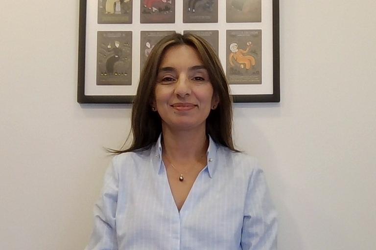 A woman with straight brown hair wearing a light blue shirt sits in front of a white wall with a framed artwork featuring several illustrations.
