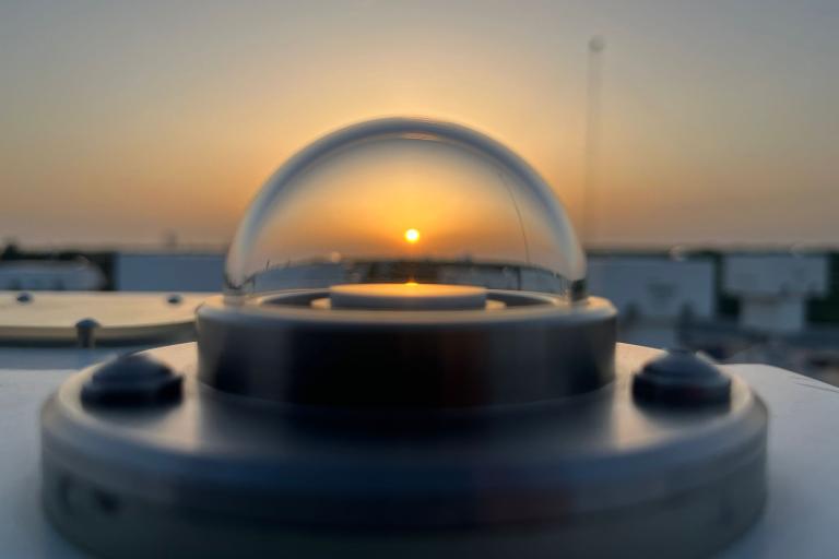 : Close-up of an atmospheric monitoring instrument at sunset, with the sun visible through its transparent dome.