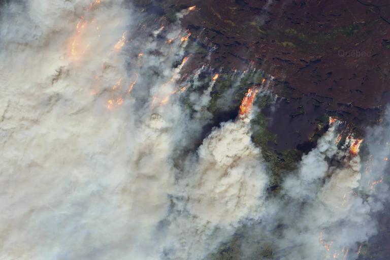 A large wildfire burns through a forested area, producing thick plumes of smoke and visible flames, as seen from an aerial or satellite perspective.