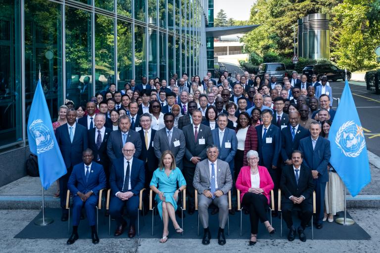 A large group of formally dressed people pose for a group photo outside a modern building, flanked by two United Nations flags.