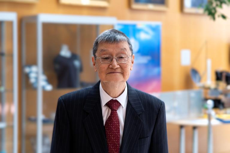 An elderly man in a dark pinstripe suit, white shirt, and red tie stands indoors with display cases and framed pictures in the background.