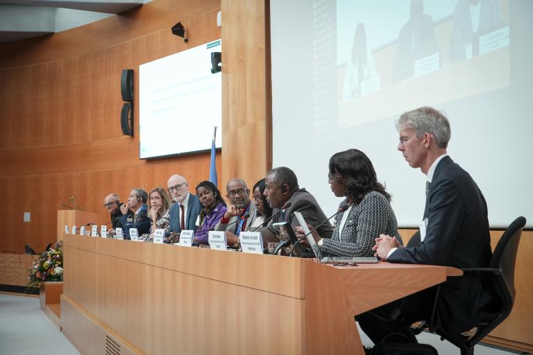 A panel of speakers sits at a long table in a conference room, participating in a formal discussion with microphones and nameplates in front of them.