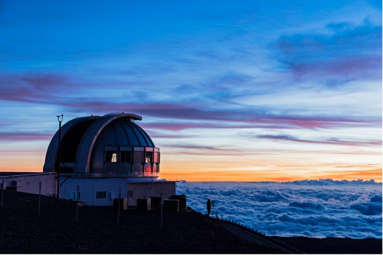 A large observatory dome sits above a sea of clouds at sunset, with a person standing nearby on a mountain slope.