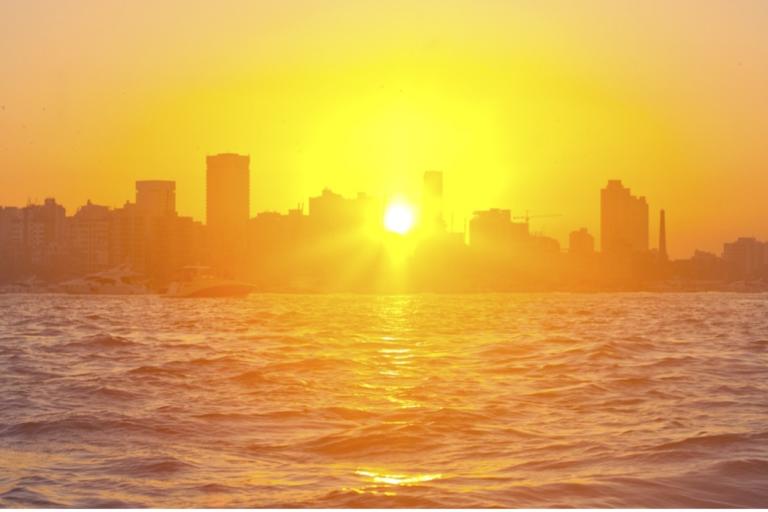 A bright orange sunset over a city skyline, with the sun low above the horizon and water in the foreground.