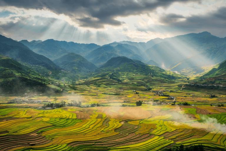 Sunlight streams through clouds over terraced rice fields and green mountains, creating a patchwork landscape in a rural valley.
