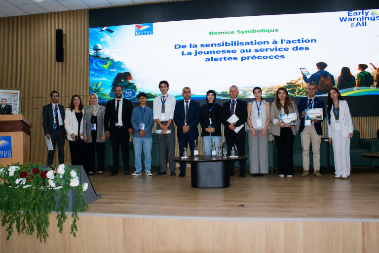 A group of people in business attire stand on stage in front of a screen displaying a French presentation about youth involvement in early warning systems.