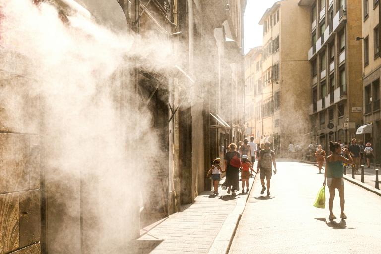 People walk down a sunlit street as mist or steam sprays from a building on the left, creating a hazy effect in the air.